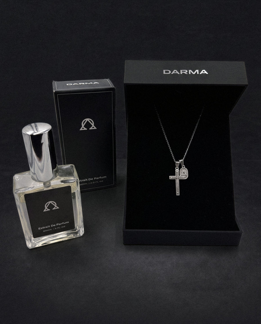 The Occasion Set in 925 Silver: the Pax Cross in 925 sterling silver in its open DARMA box, beside the DARMA extrait de parfum and its box, on a dark backdrop.
