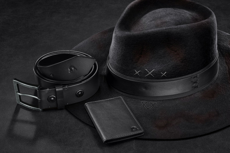 A leather belt, bifold wallet and wool felt hat in black on a dark studio sweep.