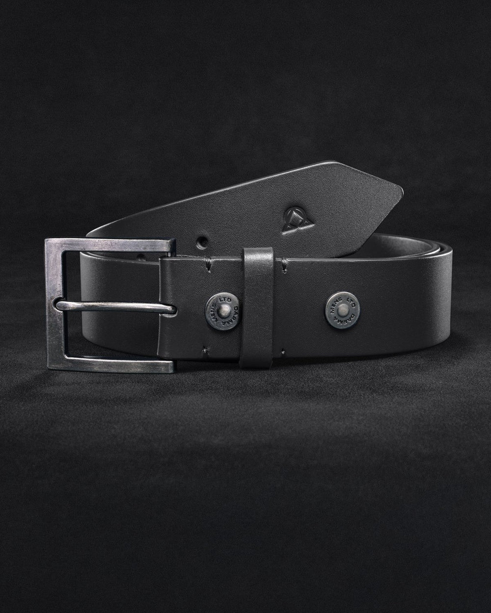 The Virtus Belt coiled on a dark textured ground, the square buckle facing the camera, two snap rivets, and the tail folded over the top showing the Alpha-Omega mark debossed. Shown in the colorway chosen on the switch below.