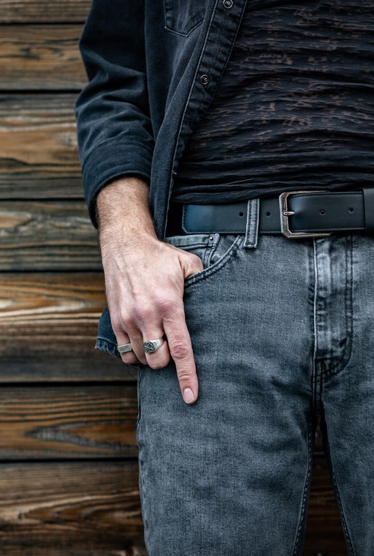 Black leather belt with a silver buckle worn with grey jeans and a black shirt, silver rings on the hand.