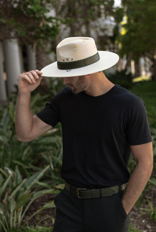 Man tipping a cream wide-brim hat with a Cypress band, black tee and Cypress belt, palms behind.