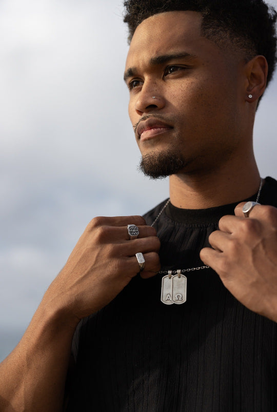 Man adjusting a silver dog tag necklace, silver rings on both hands, open sky behind.