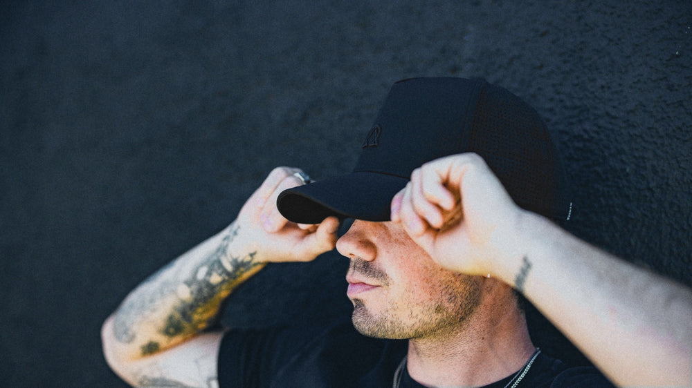 Man in a black trucker hat pulling the brim down with both hands.