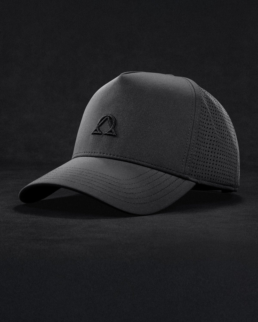 The Lux Trucker cap, three-quarter front view on a dark textured ground, the Alpha-Omega mark embroidered tonal on the front panel and the laser-perforated side panel in low light. Shown in the colorway chosen on the switch below.