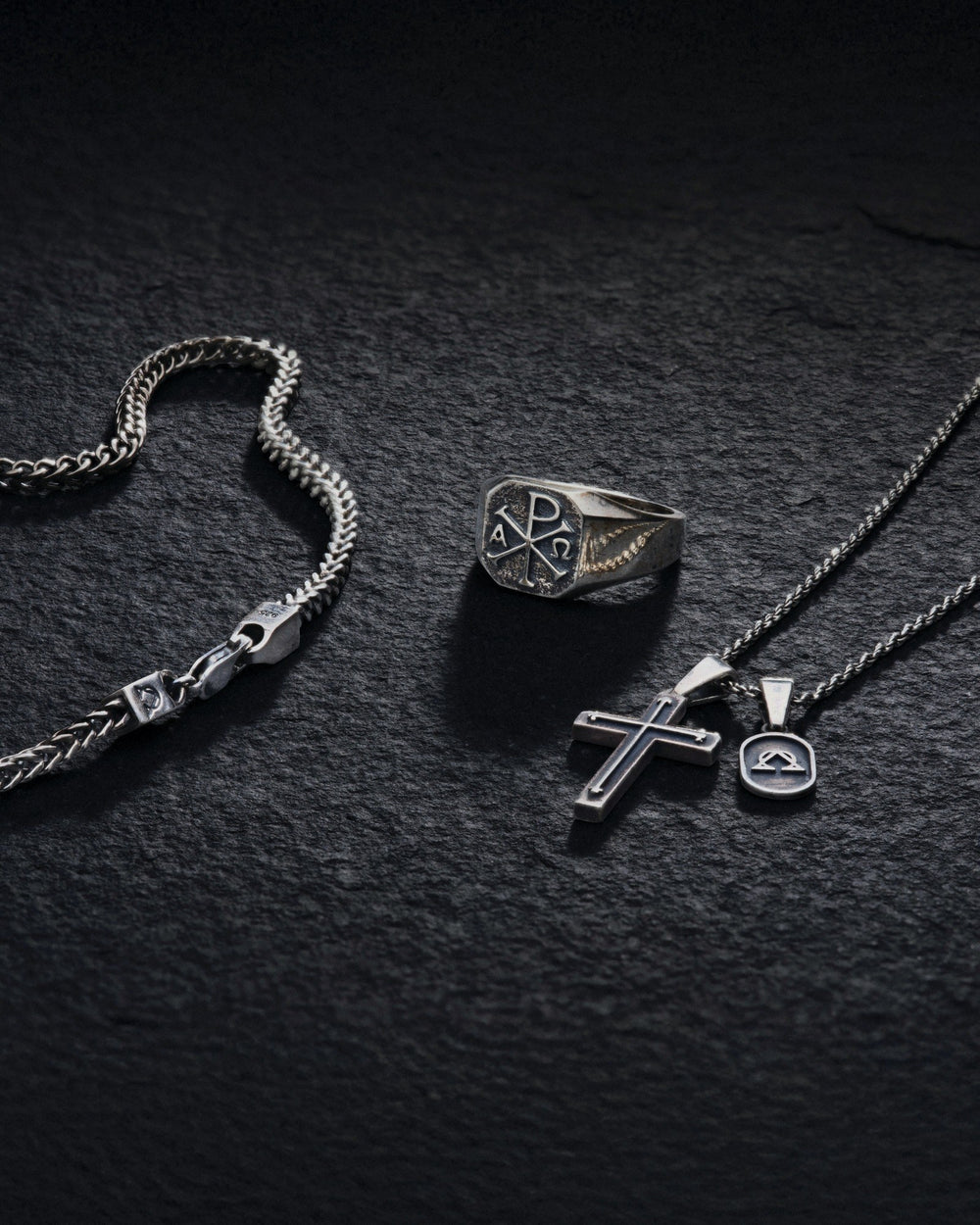A 925 sterling silver flatlay on dark slate: a franco chain curving in from the left, the Chi-Rho signet ring at centre, and a cross pendant with a small Alpha-Omega tag on a fine chain at right.