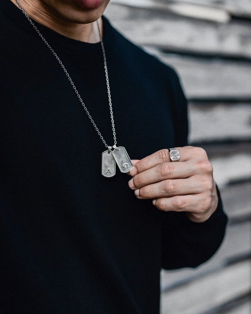 Primis Finis dog tags in 925 sterling silver worn over a black crew neck, held between the fingers, a signet ring on the hand.