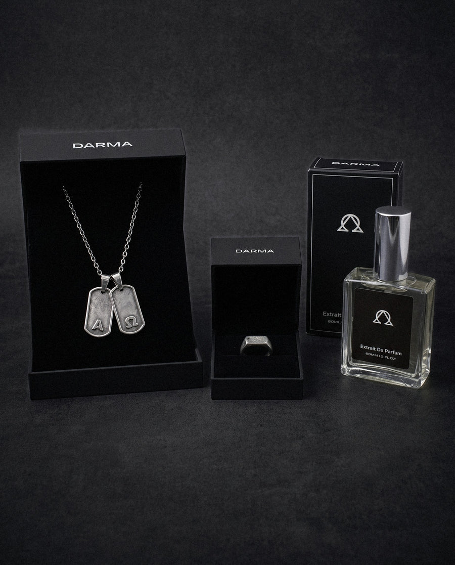 The Oath in 925 Silver: the Primis Finis dog tags and the Votum ring in 925 sterling silver in open DARMA boxes, with the DARMA extrait de parfum and its box, on a dark backdrop.