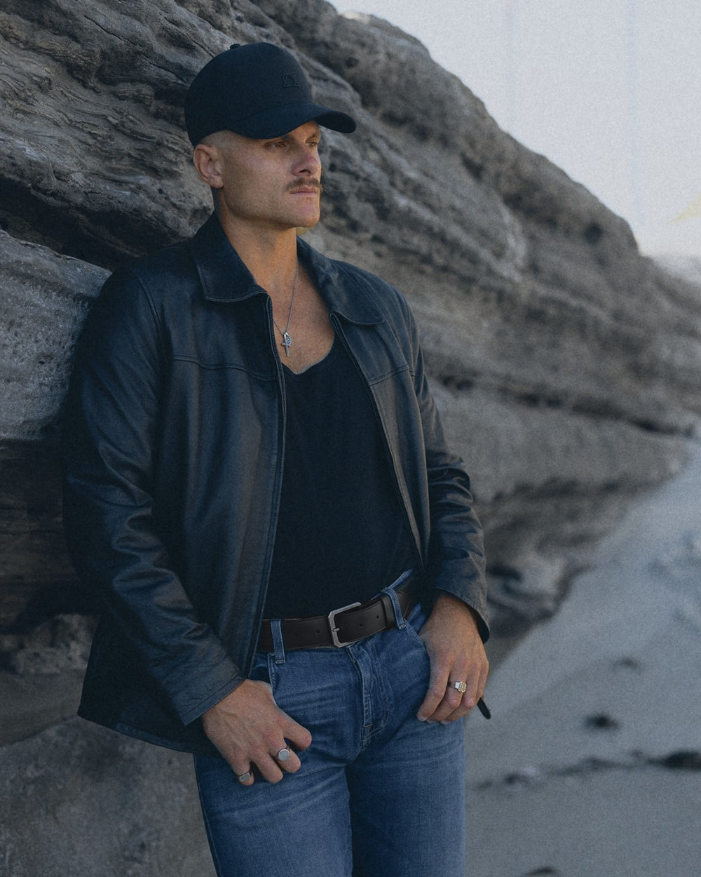 Man in a black leather jacket leaning on a sea cliff, black leather belt over blue jeans, silver rings on his hands.