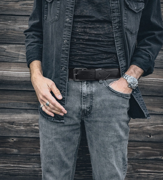 Black leather belt with a silver buckle worn with grey jeans and an open black denim shirt, wood wall behind.