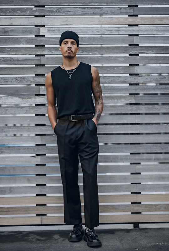 Man standing against a slatted wall in a black beanie, black tank and black trousers with a black leather belt.