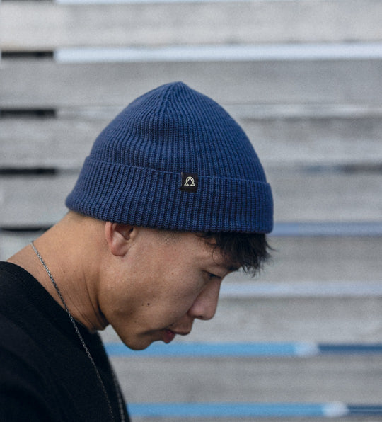 Man in profile wearing a navy knit beanie, head bowed, slatted wall behind.