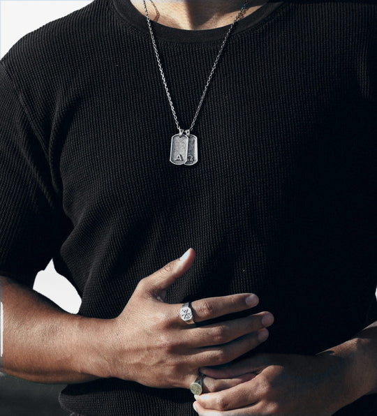 Silver dog tag necklace over a black knit tee, silver signet rings on both hands.