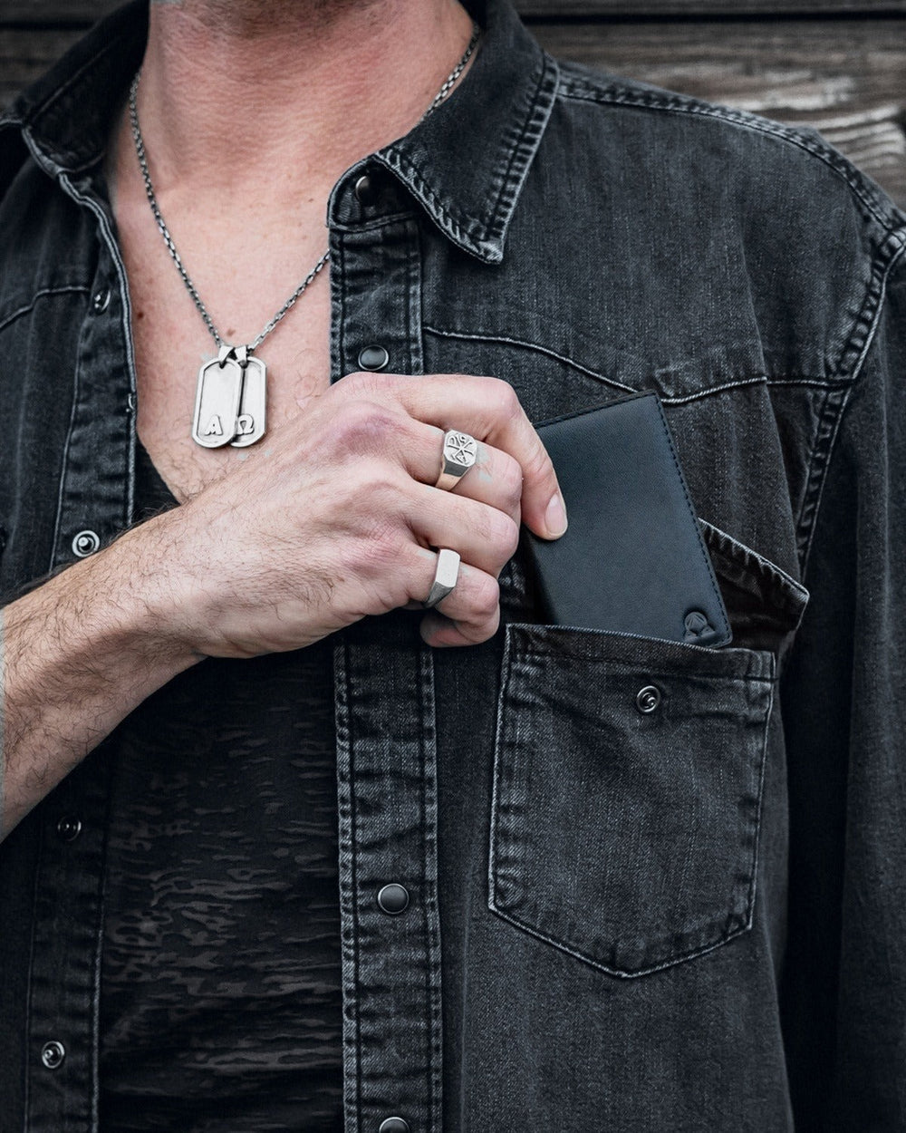 Onyx full-grain leather wallet going into the chest pocket of a black denim shirt, silver dog tags and rings, weathered wood wall behind.