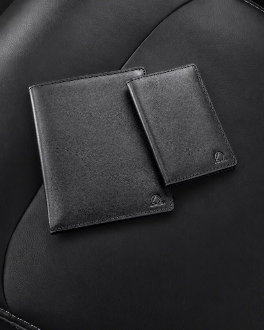 Two full-grain leather bifold wallets overlapping on a black leather car seat, shot from above at a slight angle, the Alpha-Omega mark embossed in the lower corner of each. Shown in the colorway chosen on the switch below.